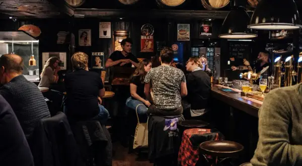 Gente en un bar oscuro, con barriles en el techo. Se preguntan cuanto gana un bar al mes mientras beben cerveza.