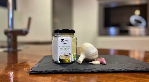 Tarro de alioli casero ecológico junto a una cabeza de ajo y dientes. ¡Este alioli se puede congelar para disfrutarlo más tiempo!