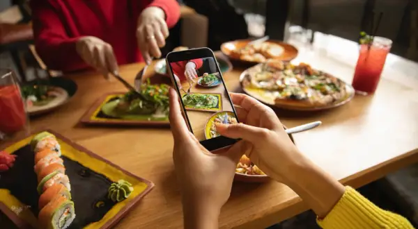 Persona toma foto de su comida en un restaurante para compartir en redes sociales. Se ven sushi, pizza y ensalada.