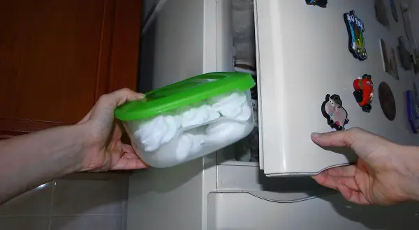 Manos guardan un recipiente con merengues blancos en el congelador. Así se puede congelar el merengue para conservarlo.