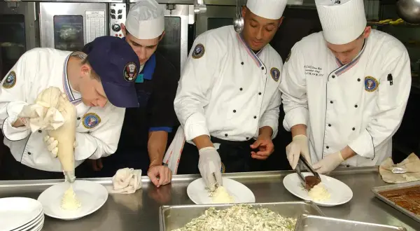 Un ayudante de cocina usa una manga pastelera para decorar un plato. Otros cocineros preparan la comida en una cocina profesional.