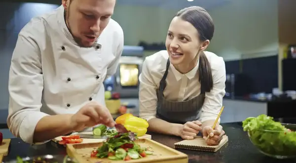 Chef instructor muestra cómo emplatar una ensalada a una alumna atenta en cursos de chef.