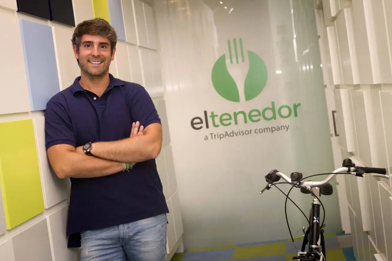 Hombre sonriente apoyado en pared colorida, con logo de El Tenedor, empresa de TripAdvisor, y una bicicleta.
