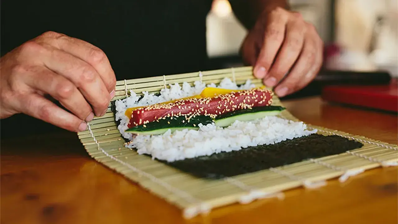 Manos expertas demuestran como se enrolla el sushi, colocando atún, pepino y pimiento sobre arroz y alga nori en una esterilla de bambú.
