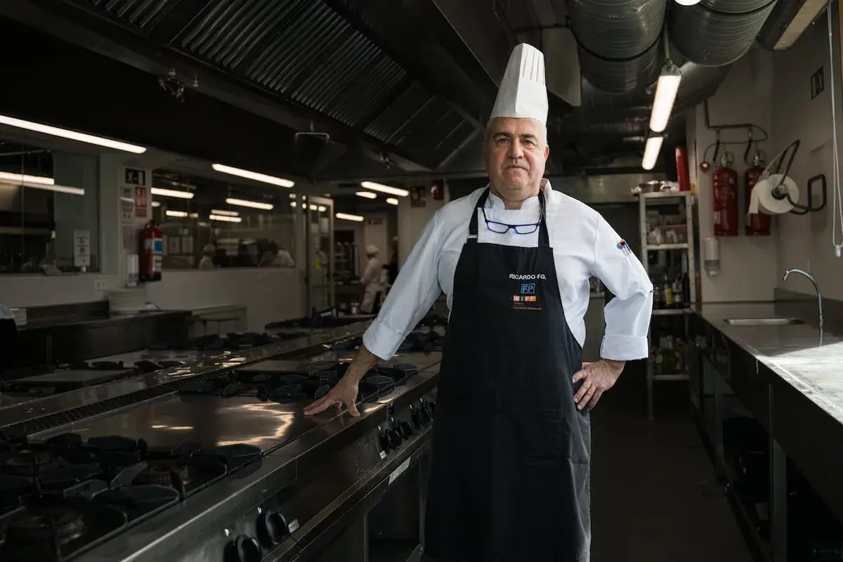 Un cocinero con gorro blanco y delantal negro posa en una cocina profesional.