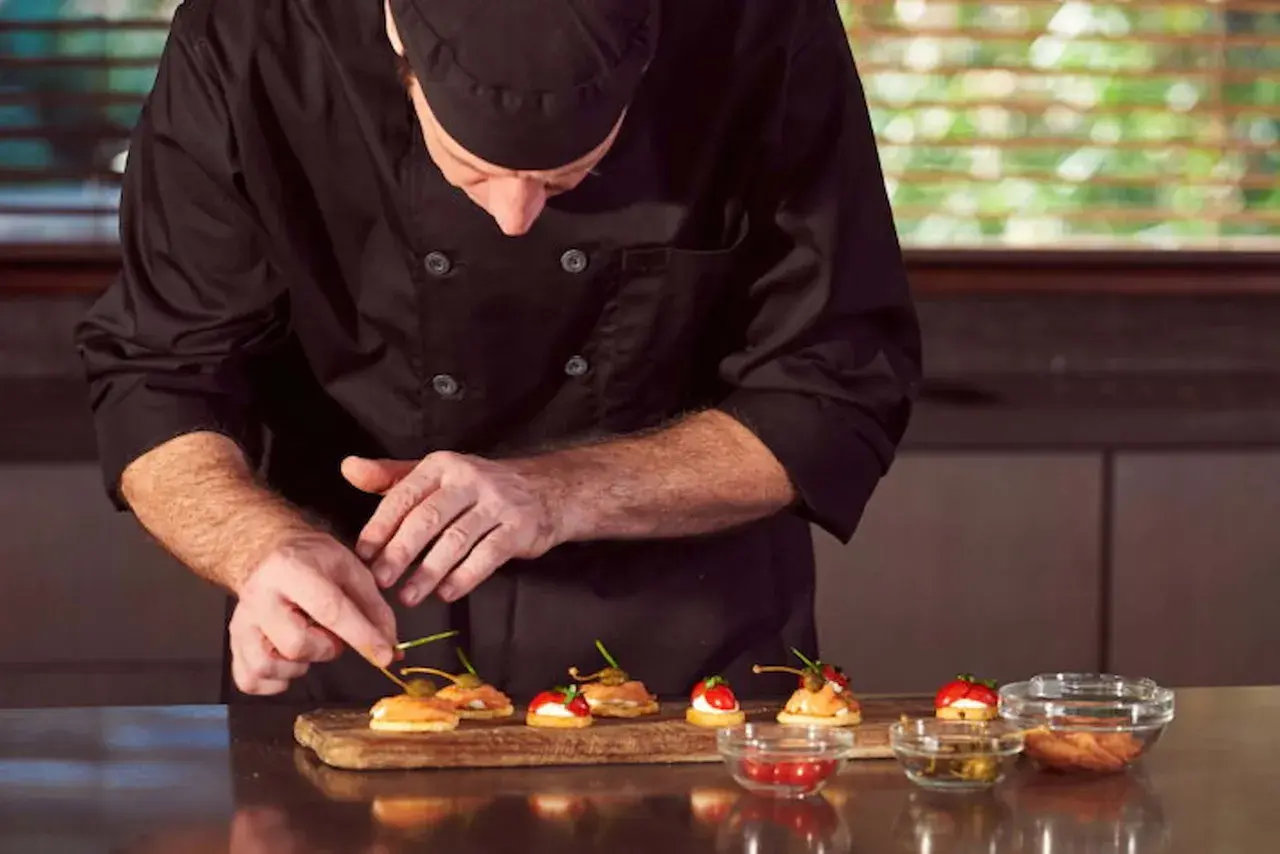 Chef en uniforme negro prepara canapés con salmón y alcaparras. Cada detalle cuenta en este arte de metre de cocina.