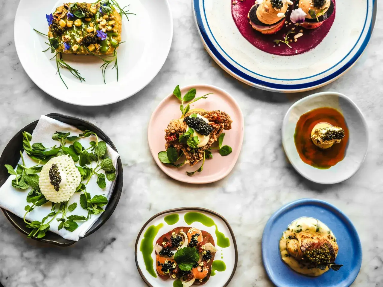 Deliciosos platos de un menú degustación, con caviar, flores comestibles y salsas vibrantes, presentados sobre una mesa de mármol.