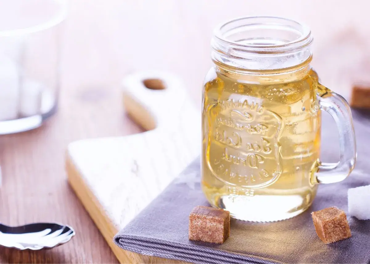 Jar de vidrio con líquido dorado, ideal para tu azucar invertida receta. Cubos de azúcar y tabla de madera al fondo.
