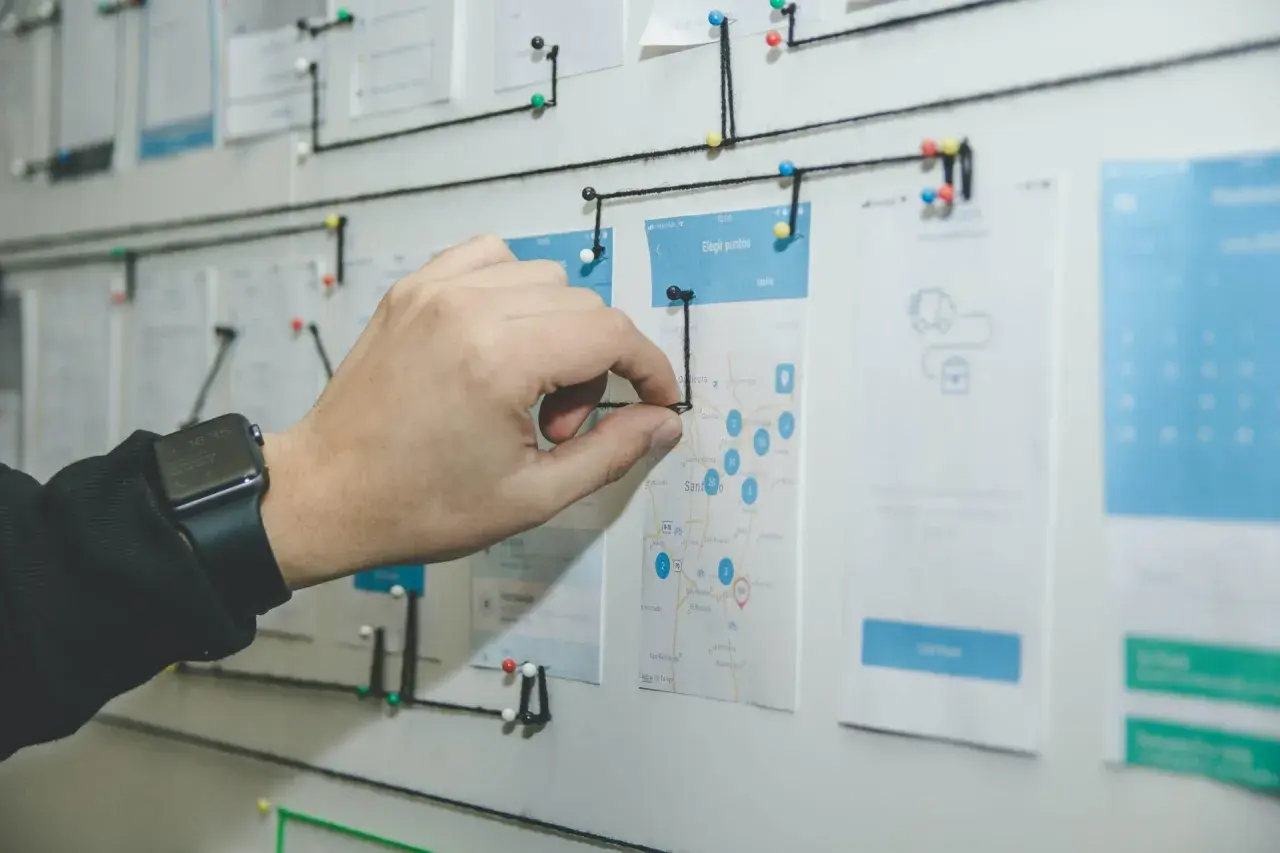 Un tablero visual con un mapa y wireframes de app, conectado con hilos. Una mano ajusta un hilo, demostrando la organización y flujo de trabajo que busca el **lean management**.