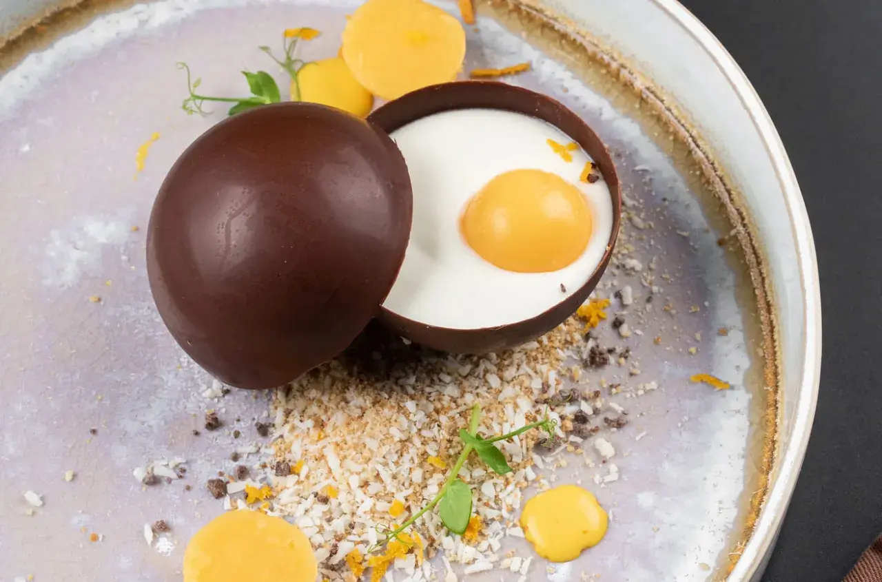 Delicioso trampantojo de comida: una esfera de chocolate oscuro abierta revela un postre cremoso con una yema simulada, rodeado de coco rallado y detalles naranjas.