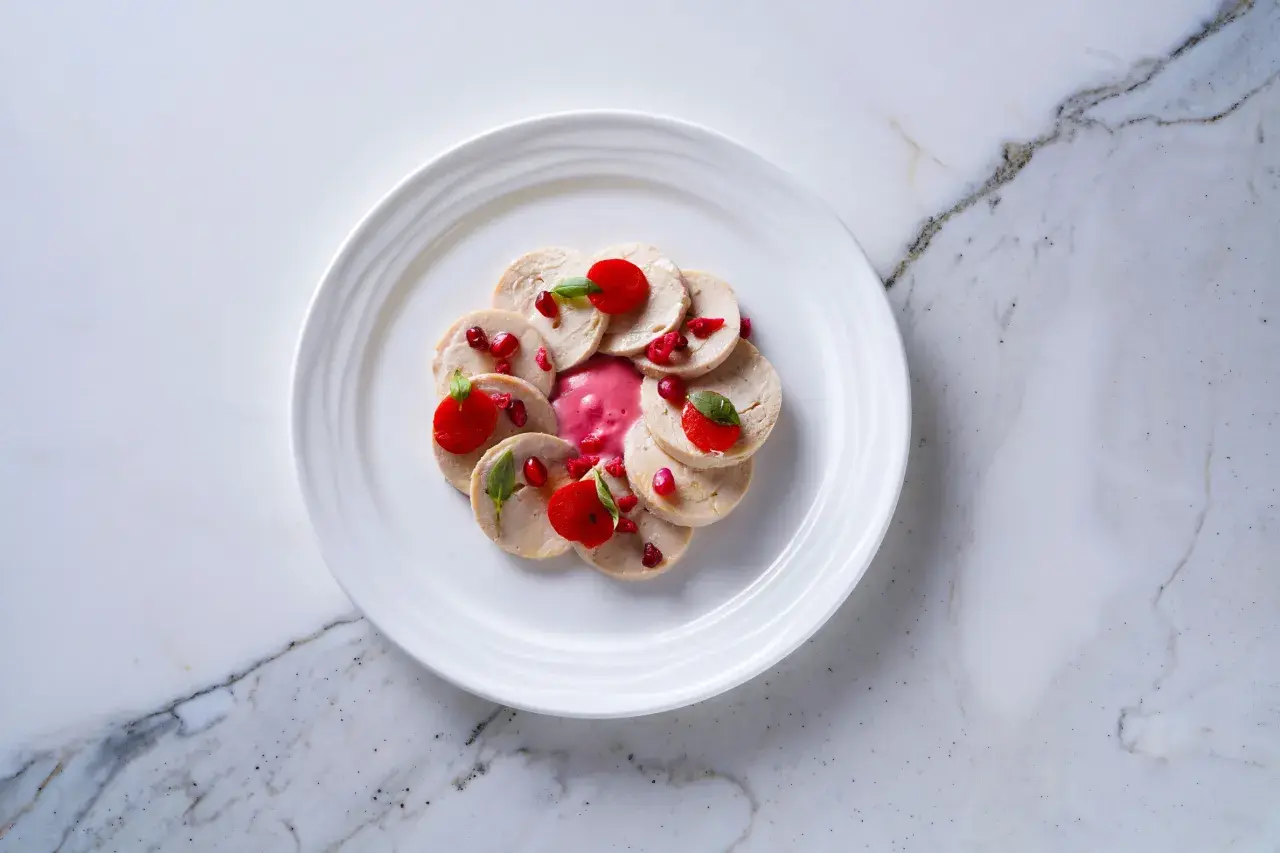 Un delicado plato estrella michelin: finas lonchas de pollo con salsa rosa, granada y hojas verdes, presentado en un plato blanco sobre mármol.