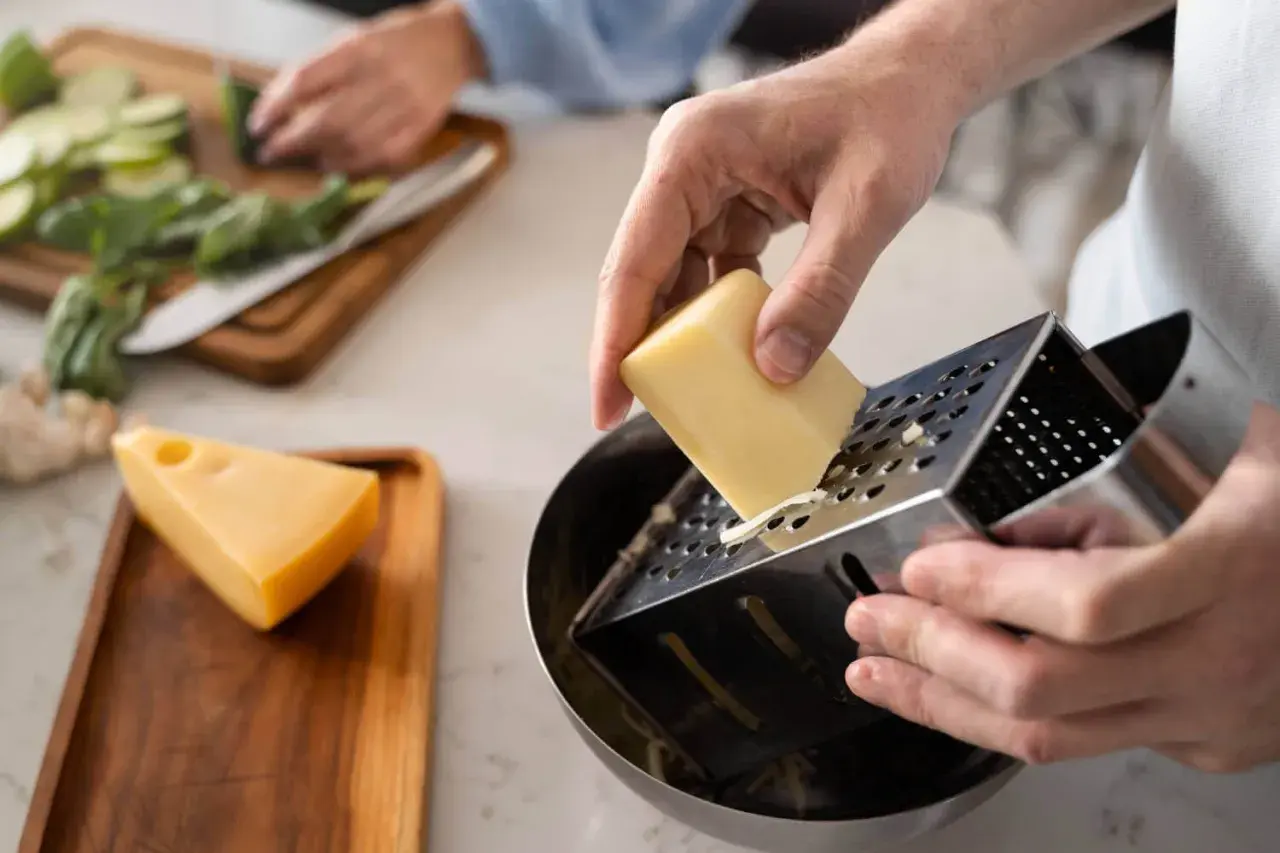 Manos rallando queso para una deliciosa comida. En el fondo, se preparan verduras.