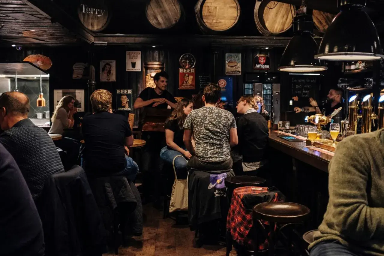 Gente en un bar oscuro, con barriles en el techo. Se preguntan cuanto gana un bar al mes mientras beben cerveza.