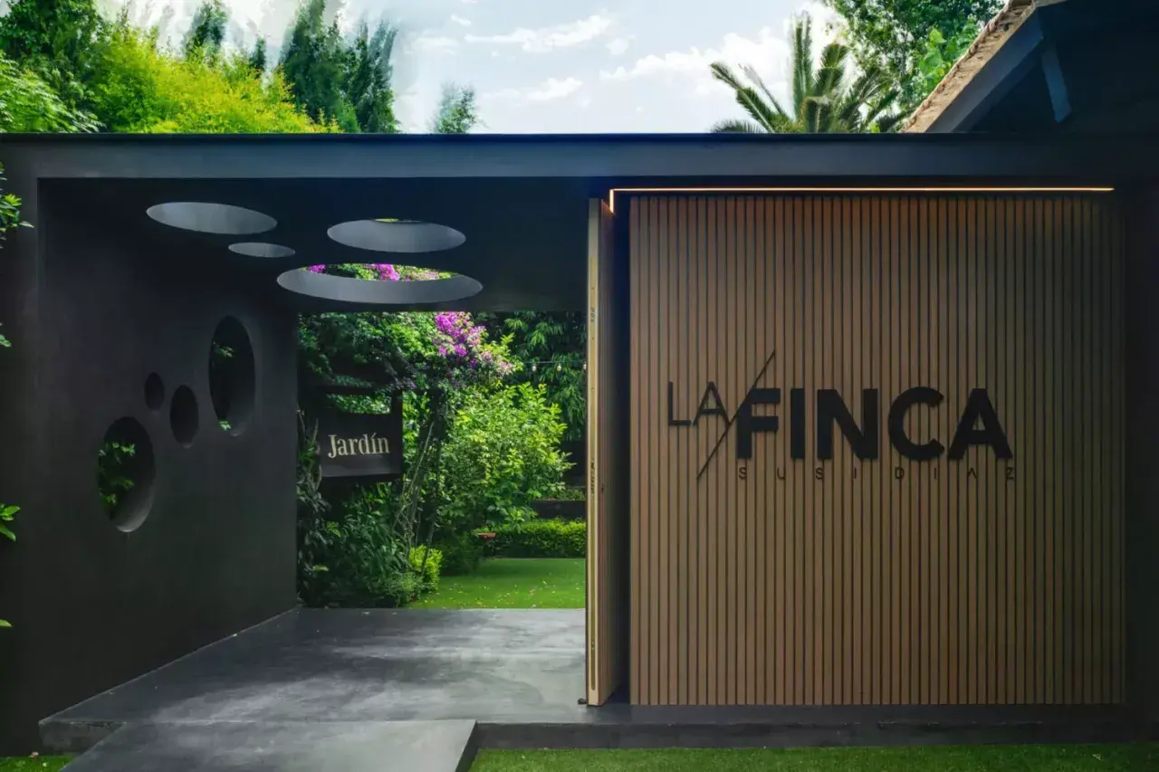 Entrada a "La Finca" de Susi Díaz, un jardín exuberante con un letrero de madera y una fachada moderna.