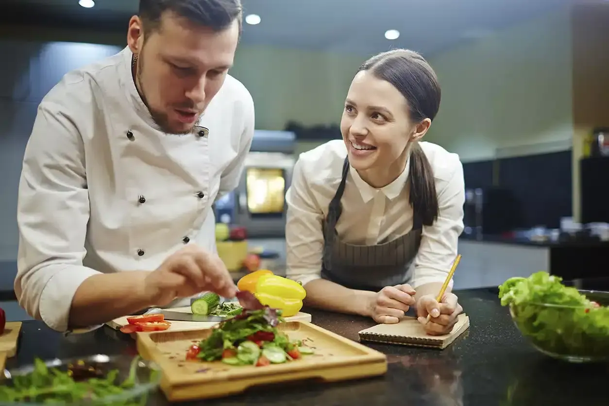 Chef instructor muestra cómo emplatar una ensalada a una alumna atenta en cursos de chef.
