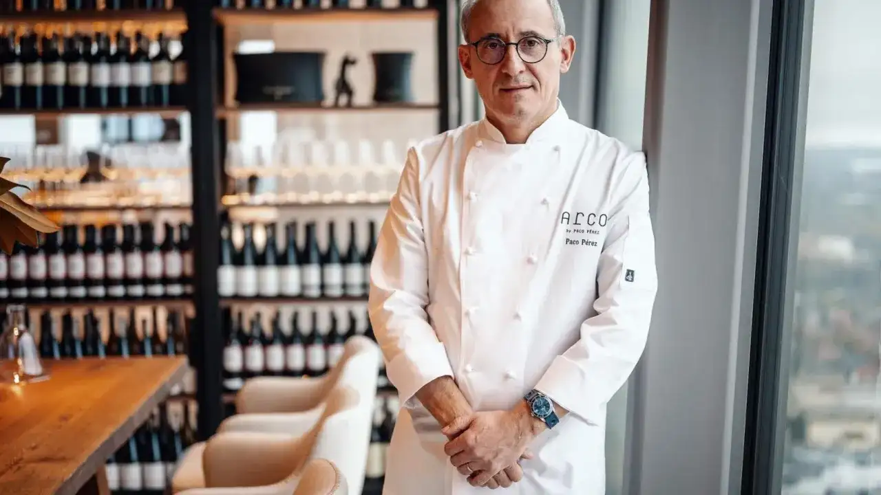 Chef Paco Pérez, con su chaquetilla blanca, posa con confianza en un restaurante elegante, rodeado de botellas de vino.