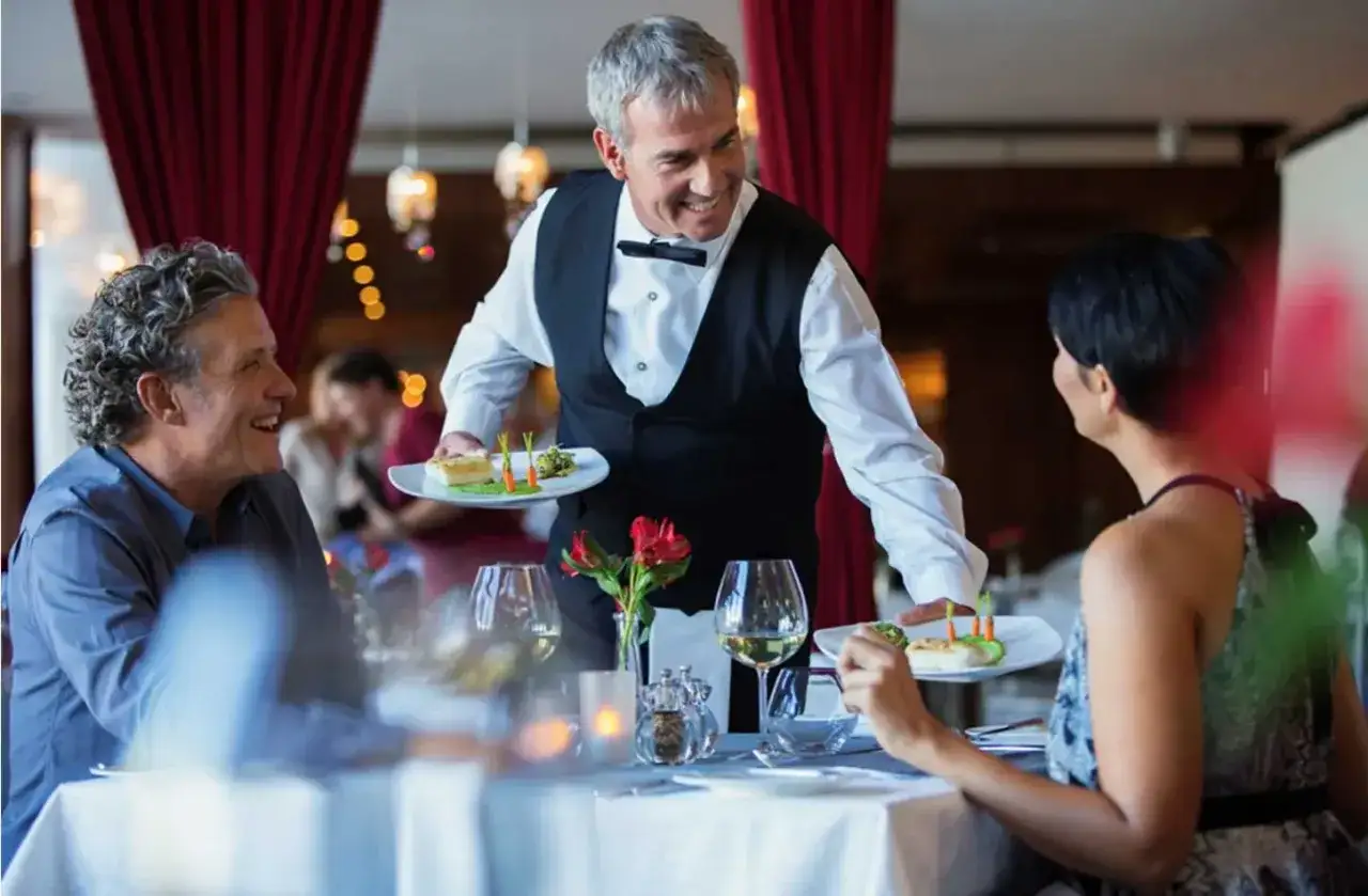 Un camarero sonriente sirve platos a una pareja en un restaurante. Un ejemplo de como ser un buen camarero.