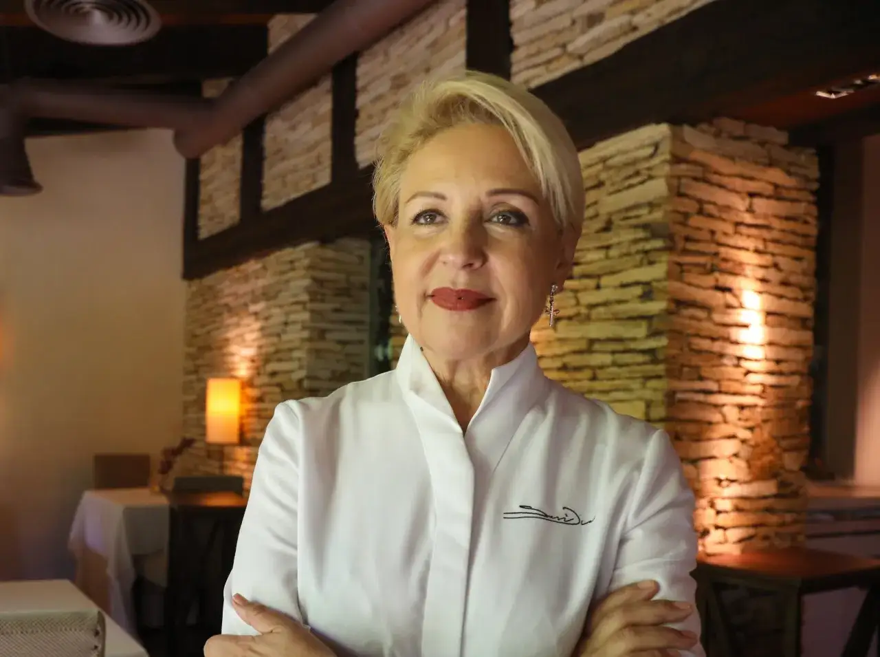 Susi D&iacute;az, chef con su distintiva firma bordada en su uniforme blanco, posa con confianza en un restaurante r&uacute;stico.