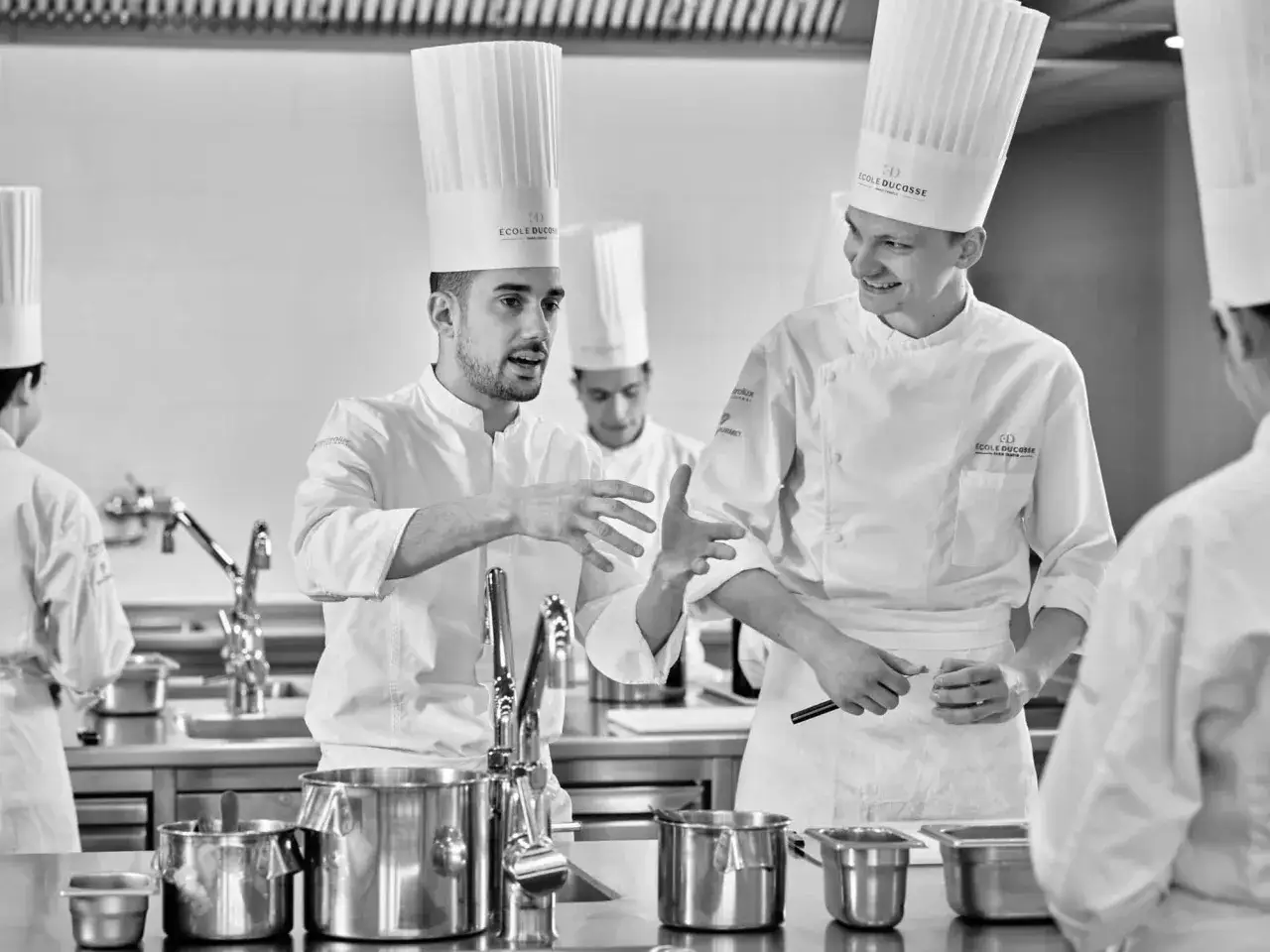 Dos chefs con gorros altos discuten t&eacute;cnicas culinarias, uno gesticula apasionadamente. Parecen estar en camino de convertirse en el mejor cocinero del mundo.