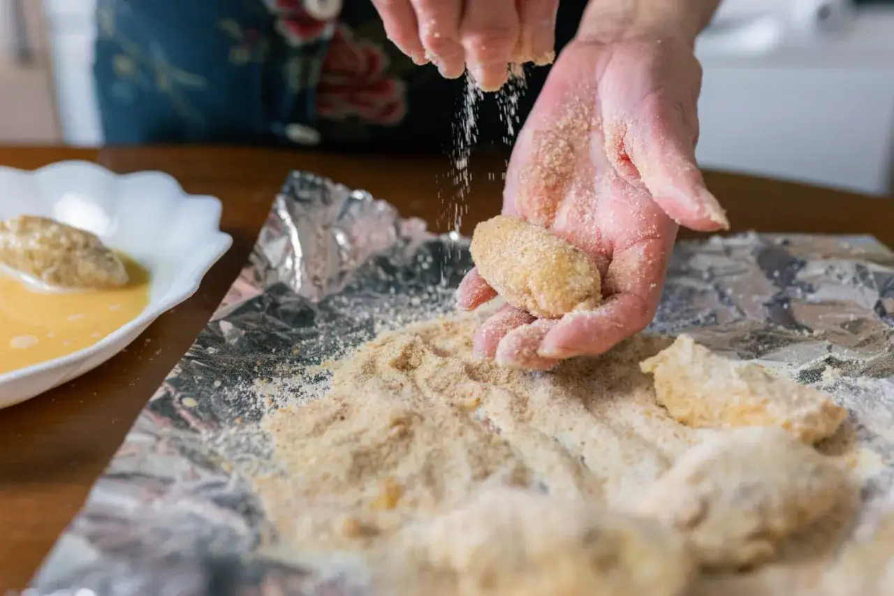 Manos cubriendo croquetas con pan rallado. La proporci&oacute;n de harina y leche para croquetas es clave para su textura.