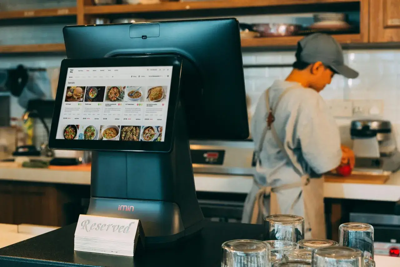 Terminal de restaurante muestra opciones de comida. Un empleado prepara algo en la cocina, quizás parte de la comida de personal.