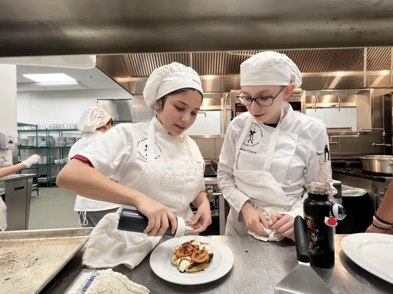 Dos estudiantes en un curso intensivo de cocina decoran un plato. Una aplica salsa a una bruschetta, la otra limpia.
