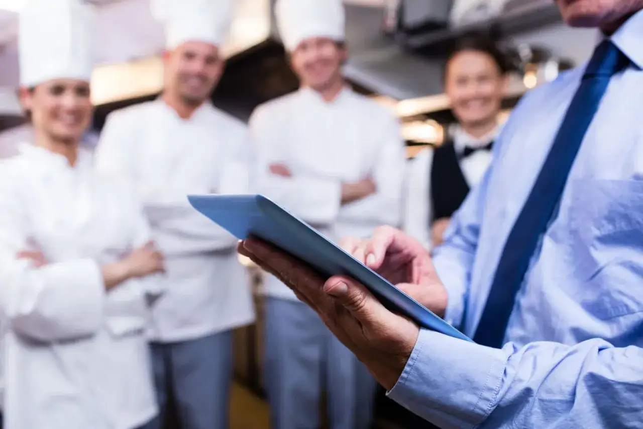 Un hombre con corbata azul revisa una tablet, mientras un equipo de cocina sonr&iacute;e al fondo. El f&b que es, se gestiona con tecnolog&iacute;a.