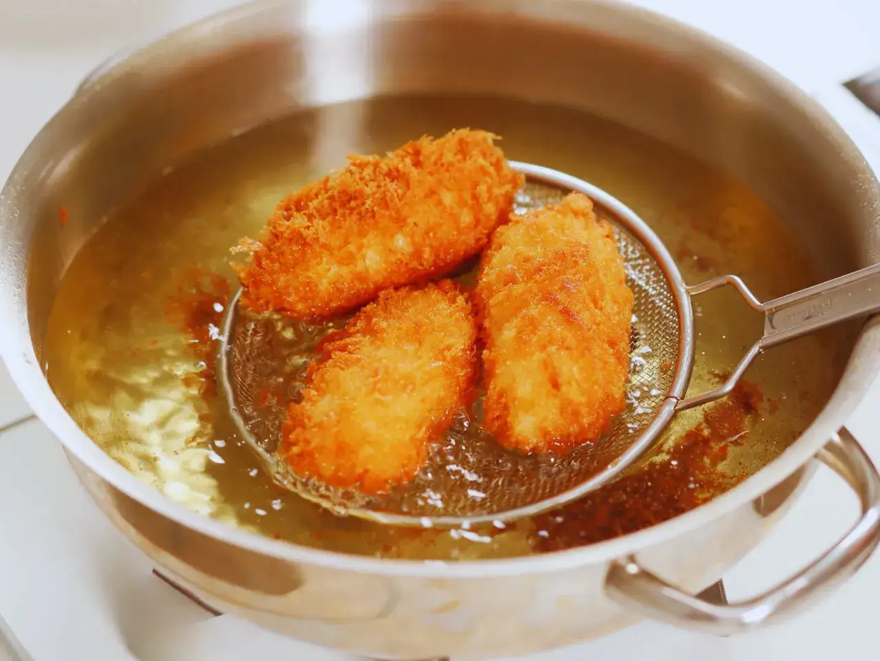 Croquetas doradas y crujientes friéndose en aceite caliente. Un colador de metal sostiene tres croquetas recién hechas, listas para disfrutar.