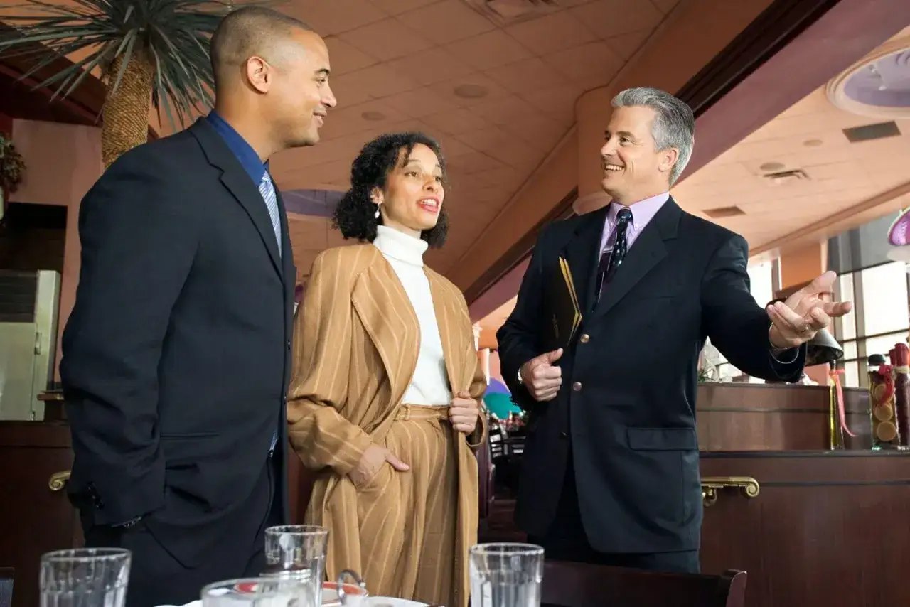 Tres personas conversan en un restaurante. Uno de ellos, con un traje marrón a rayas, parece explicar algo, quizás sobre el menú o el metre de cocina.