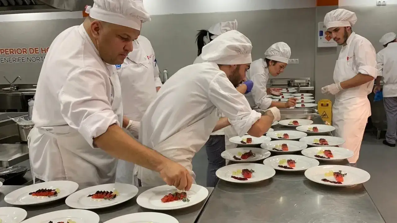 Varios cocineros con gorros blancos preparan platos con salm&oacute;n y guarniciones. Un aspirante a t&iacute;tulo de cocinero decora con esmero.