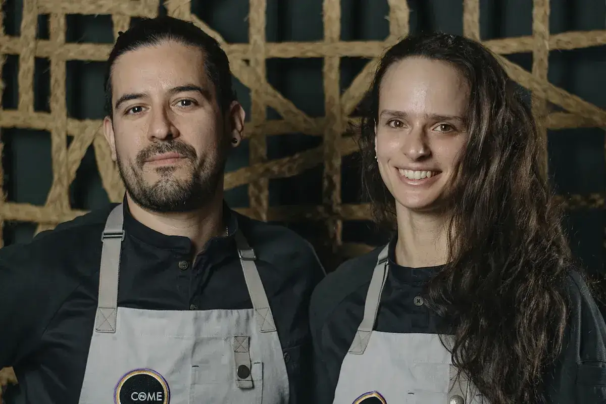 Dos chefs sonr&iacute;en, uno con coleta y barba, la otra con cabello largo. Ambos llevan delantales con el logo 