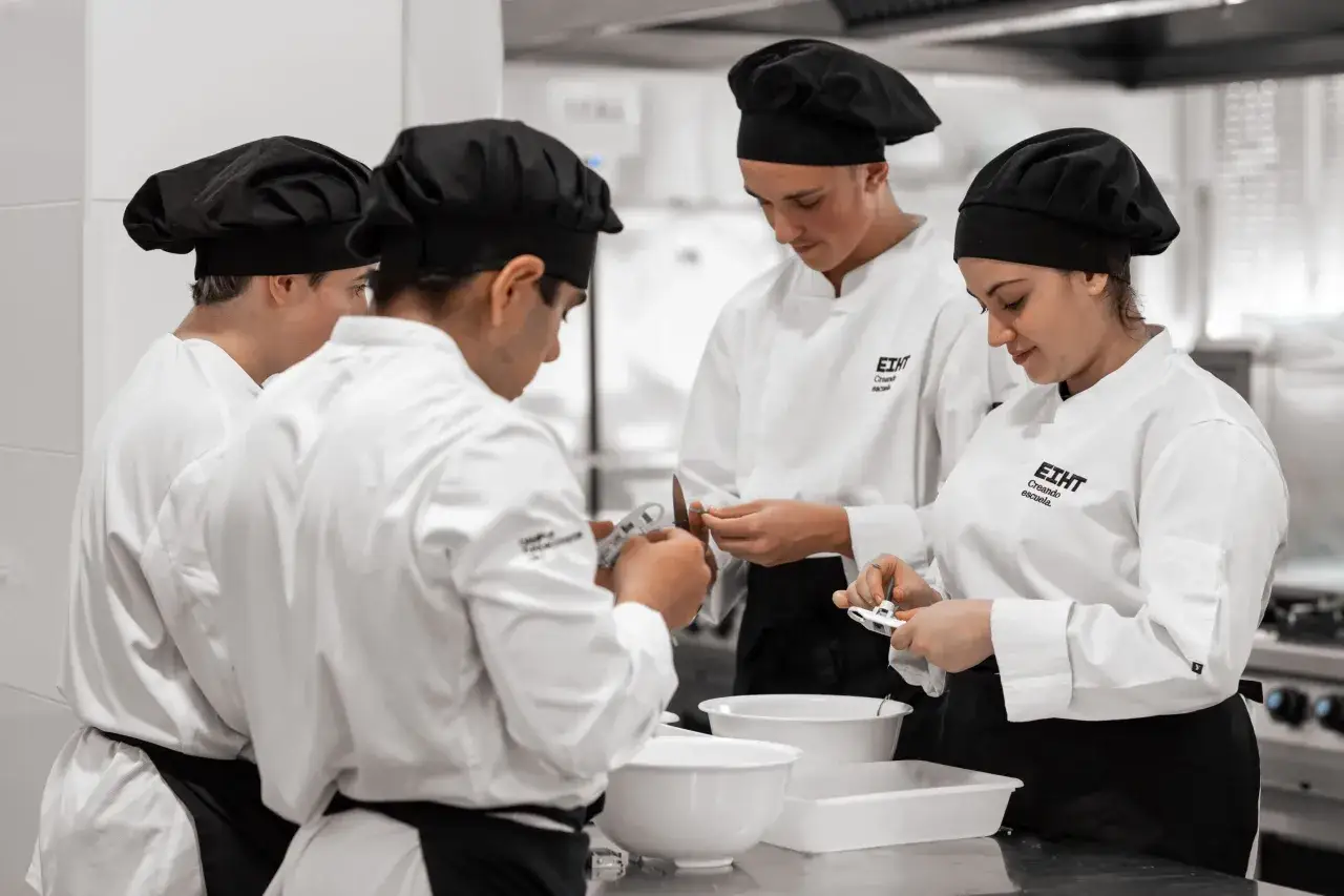 Estudiantes de cocina con chaquetillas blancas y gorros negros practican t&eacute;cnicas culinarias. Pronto obtendr&aacute;n su diploma de cocina.