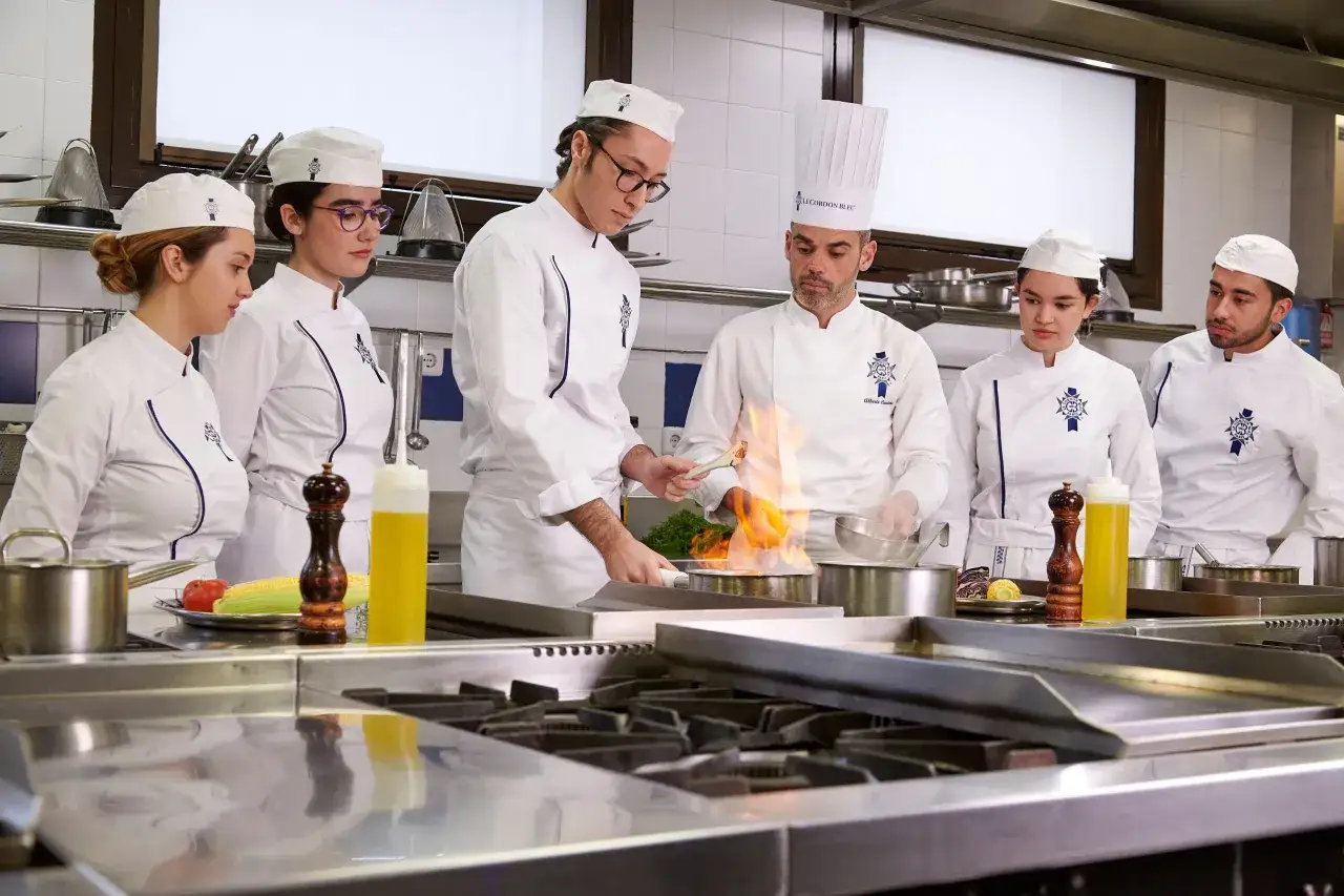 Estudiantes de grado en ciencias gastronómicas aprenden técnicas culinarias en una cocina profesional, con llamas vivas en una sartén.