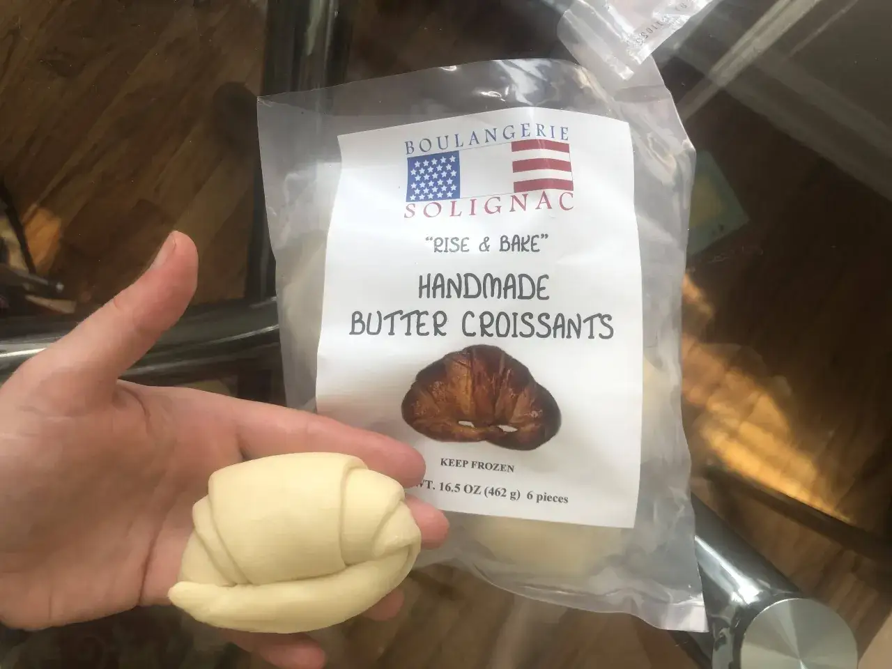Mano sosteniendo un croissant crudo junto a un paquete de croissants de mantequilla hechos a mano. &iexcl;Se pueden congelar los croissants para hornearlos m&aacute;s tarde!