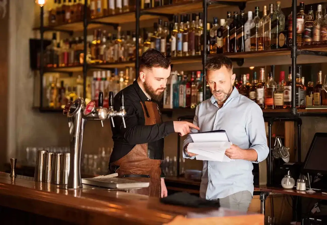 Dos hombres discuten un men&uacute; en una barra, mostrando la diferencia entre bar y restaurante. Estantes llenos de botellas de licor detr&aacute;s.