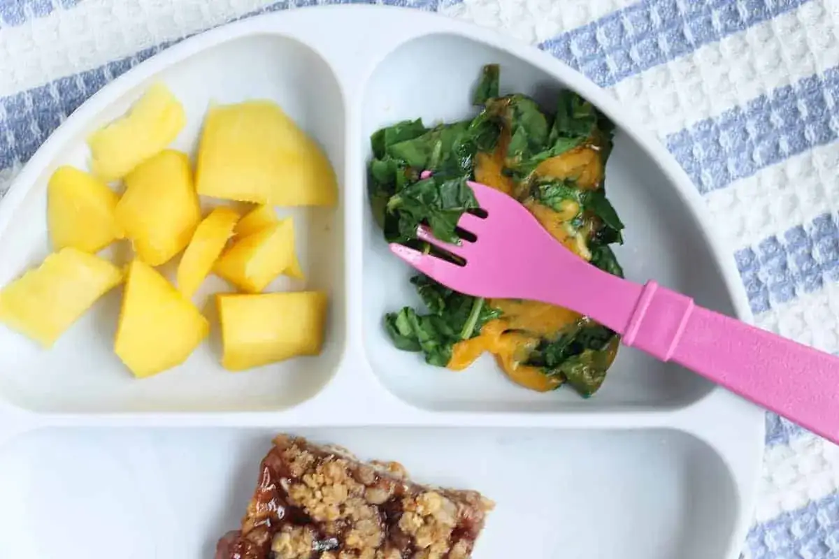 Plato dividido con trozos de mango, espinacas con queso y un trozo de barrita de cereales. &iexcl;Ideas de recetas con verduras para ni&ntilde;os!