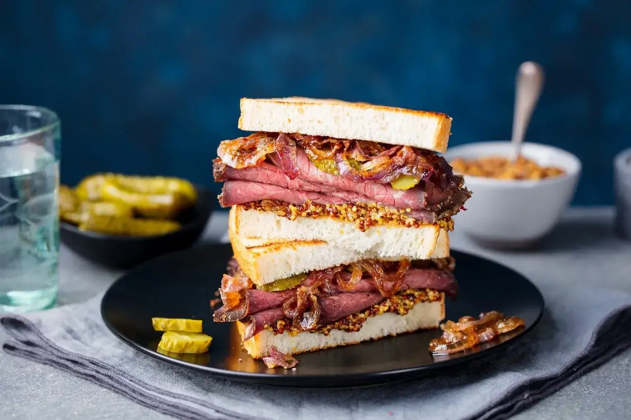 Un delicioso s&aacute;ndwich de pastrami como se come, con pan tostado, mostaza, pepinillos y cebolla caramelizada.