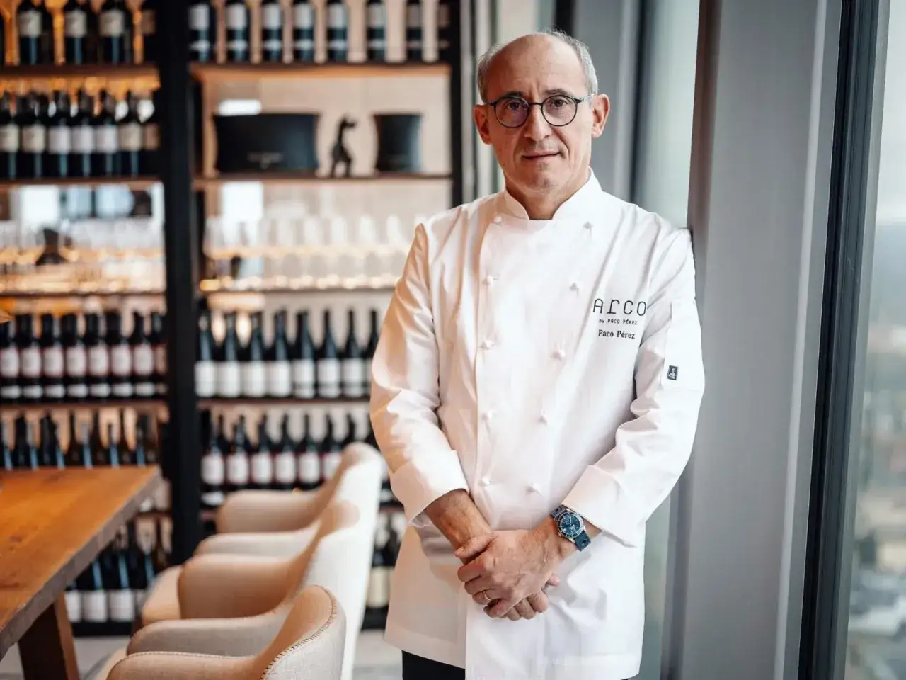 Chef Paco P&eacute;rez, con gafas y chaquetilla blanca, posa en un restaurante con estanter&iacute;as de vino.