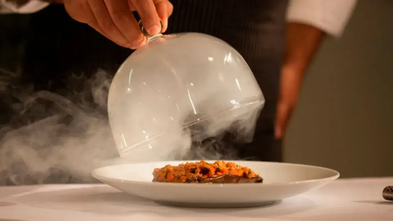 Plato de berenjena con salsa, servido bajo una campana de cristal con humo. Una experiencia culinaria digna de un bib gourmand.