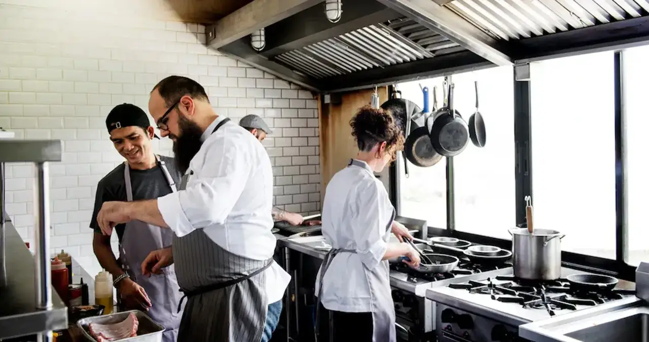 Un equipo de chefs trabaja en la cocina, mostrando el ciclo de cocina. Uno ense&ntilde;a a otro, mientras otros preparan platos.