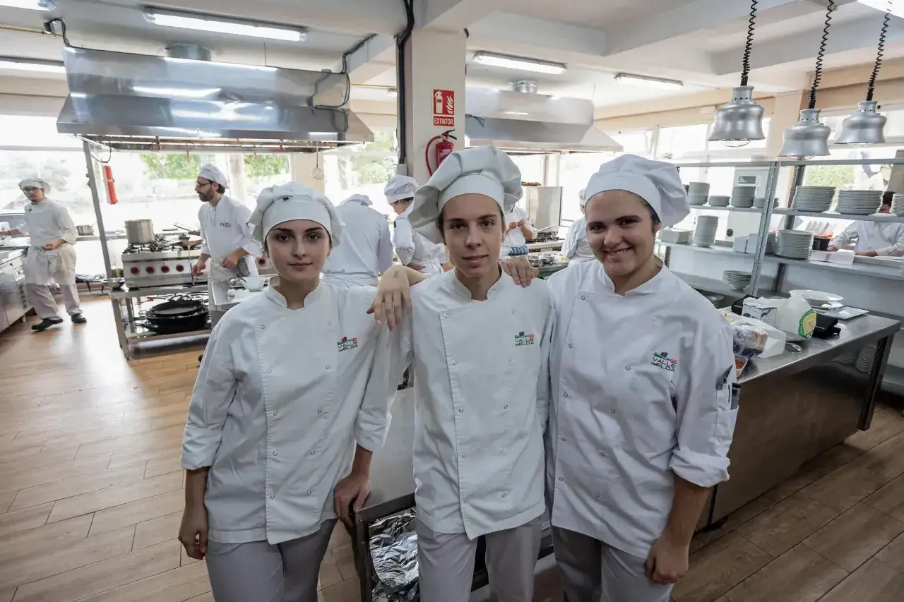 Tres jóvenes aprendices de cocina, con chaquetas blancas y gorros, sonríen en una cocina profesional. Aprenden como ser chef.