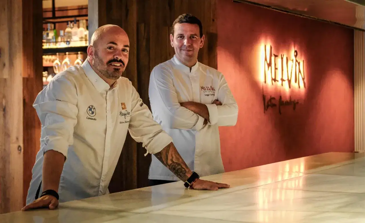 Dos cocineros famosos posan en un bar. Uno tiene un tatuaje en el brazo. Detrás de ellos, un letrero de neón dice