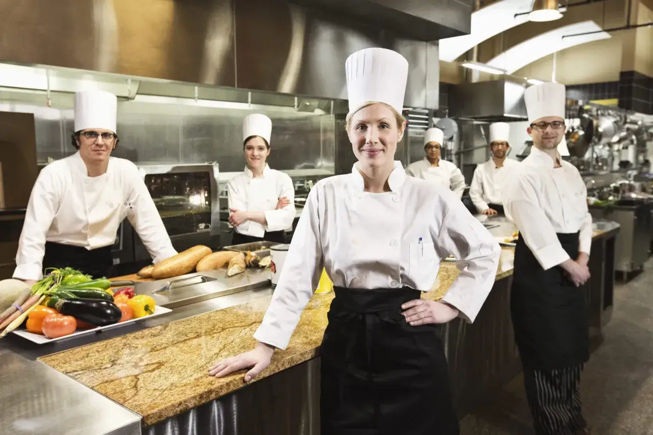 Chefs en formaci&oacute;n en cursos de cocina para profesionales, sonriendo en una cocina profesional con verduras frescas y pan en la encimera.
