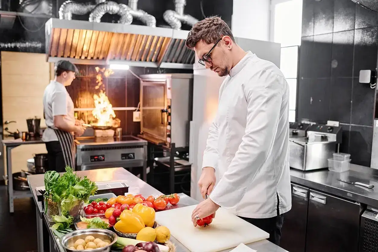 Un ayudante de cocina en España prepara ingredientes frescos mientras otro chef cocina con fuego vivo.
