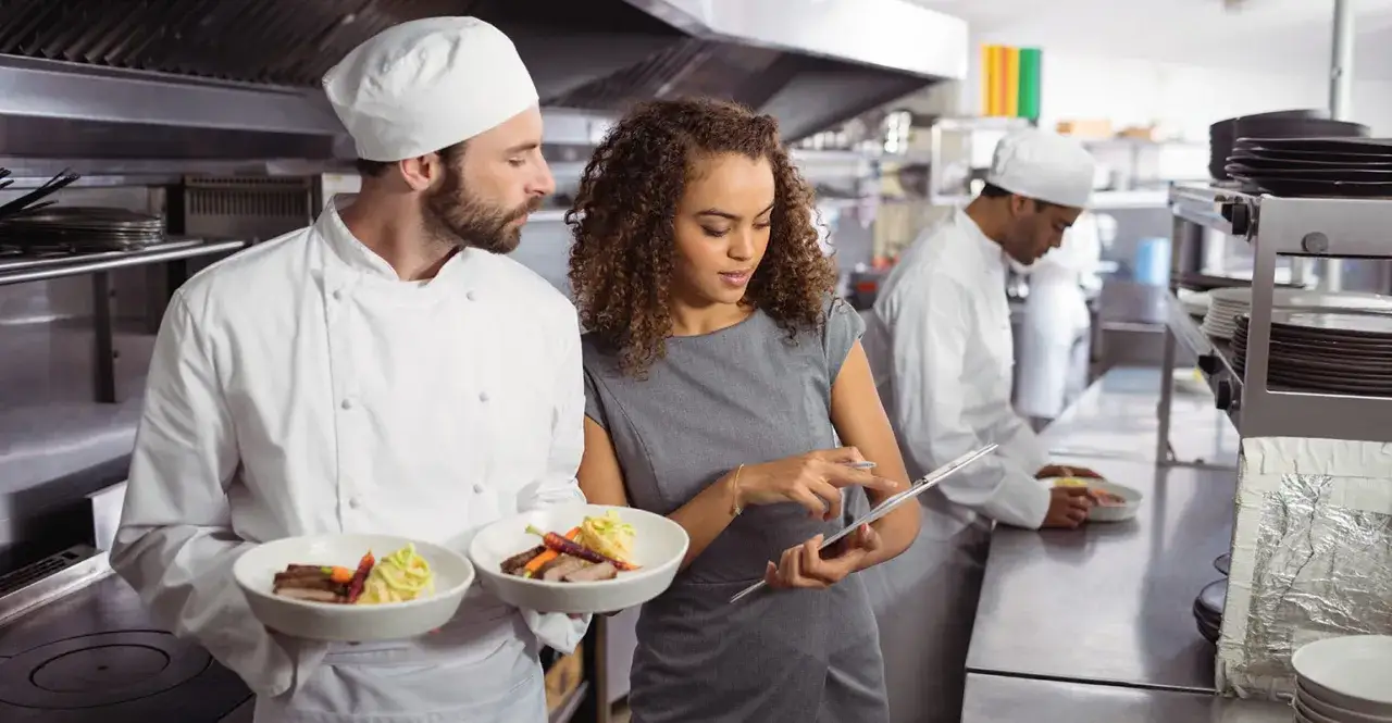 Chef y gerente revisan platos en cocina profesional. Un equipo demuestra su maestr&iacute;a en food and beverage.