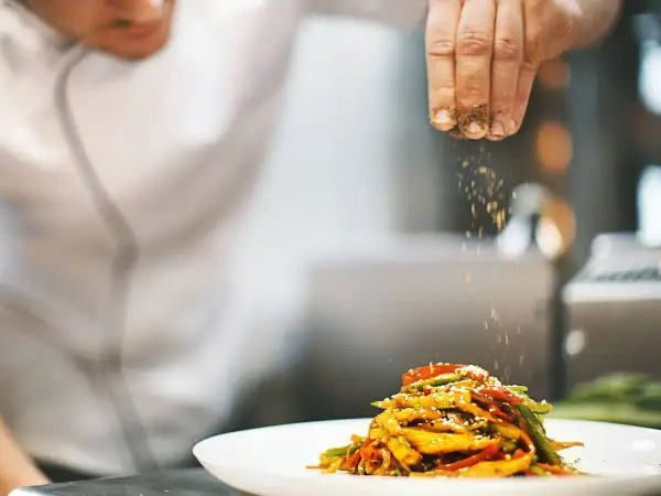 Un chef con título de cocinero espolvorea especias sobre un plato de verduras salteadas.