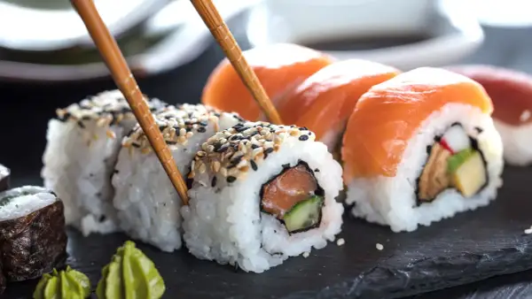 Palillos recogen un rollo de sushi con salmón y pepino, mostrando cómo se hace el sushi. Otros rollos con salmón y wasabi al lado.