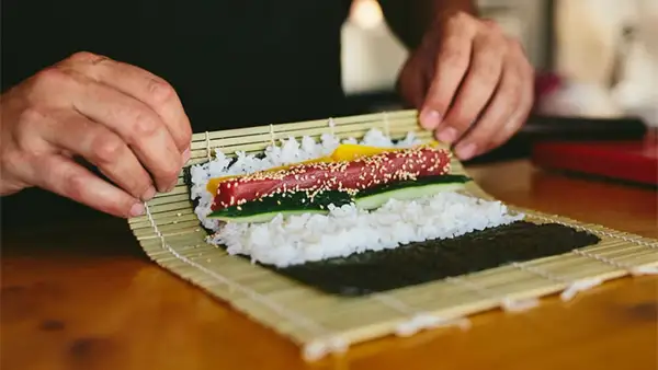 Manos expertas demuestran como se enrolla el sushi, colocando atún, pepino y pimiento sobre arroz y alga nori en una esterilla de bambú.