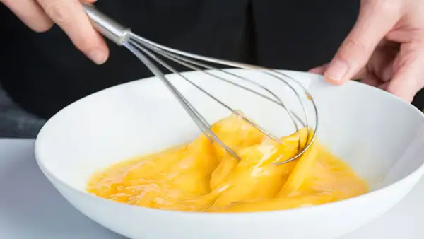 Manos batiendo huevos en un bol blanco, un paso clave para que es emulsionar y lograr texturas perfectas en la cocina.
