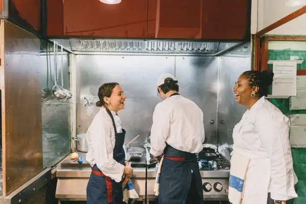 Tres chefs ríen en una cocina profesional, aprendiendo técnicas de cocina italiana.